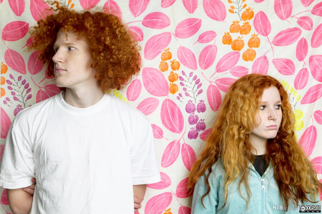 En rødhåret gutt og en rødhåret jente står inntil en vegg og ser til hver sin side. Foto.
