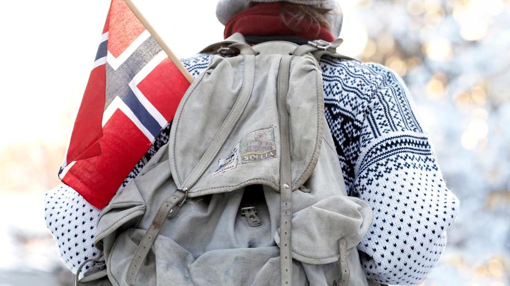 Foto av ryggen til en person med lusekofte og en gammeldags ryggsekk der det stikker opp et norsk flagg.
