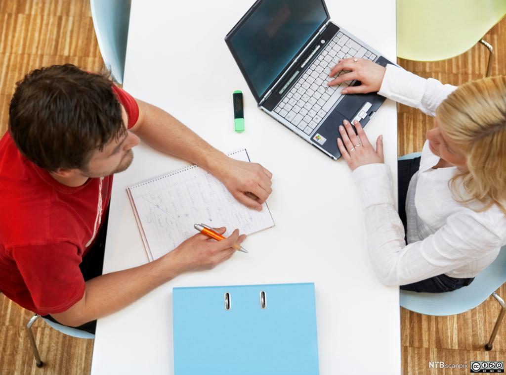 Bilde tatt ovenfra av to unge studenter, en mann og en kvinne, som sitter ved et bord med en datamaskin og  en skriveblokk. Foto.