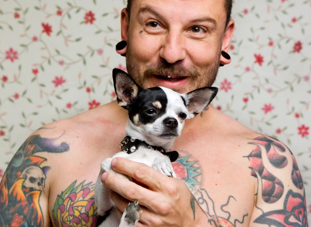Man med tavoveringer holder en liten chihuahua. Foto.