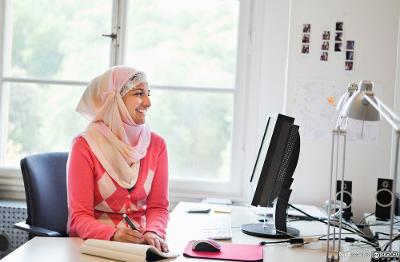 Ung jente i hijab sitter forran en datamaskin og tar notater. Foto.