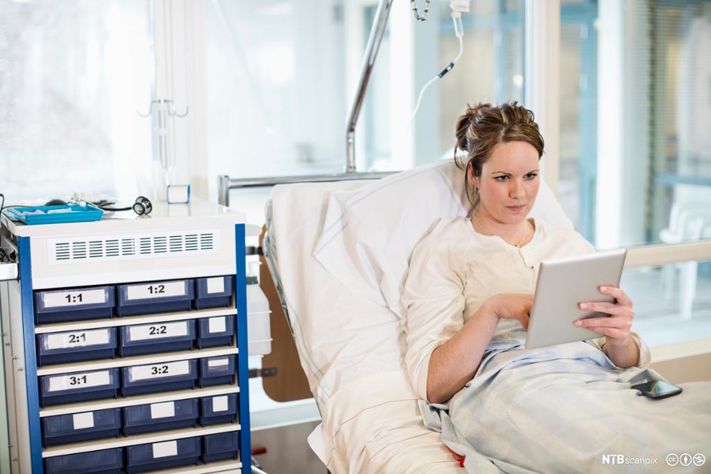 Ung kvinne leser på nettbrett mens hun ligger i sykehusseng. Foto.