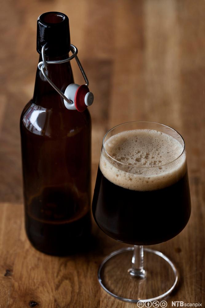 Et ølglass med mørkt øl og en ølflaske på et trebord. Foto.