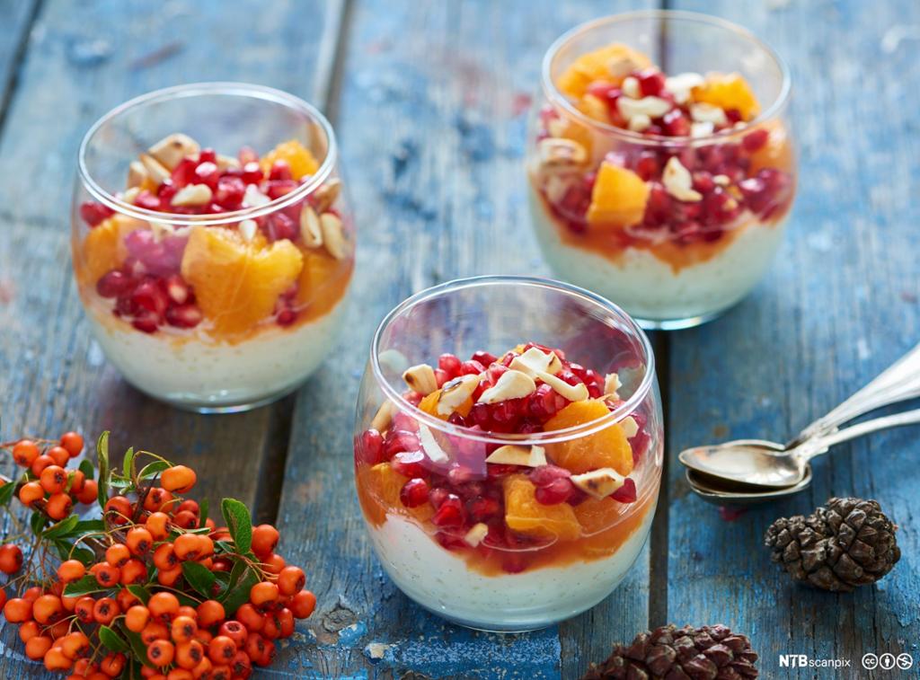 Tre runde dessertglas med kvit pudding med fruktsalat med mellom anna granateplefrø. Glasa står på grove treplankar og bordet er pynta med rognebær og kongler. Foto.