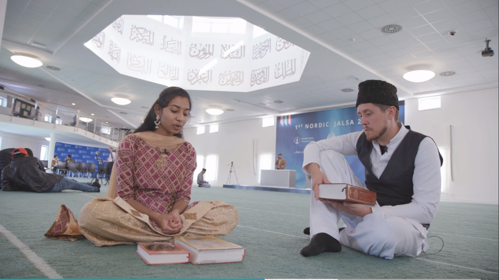Kvinne i sari og mann i tradisjonell muslimsk drakt sitter på gulvet i en moské med bøker foran seg. Foto.