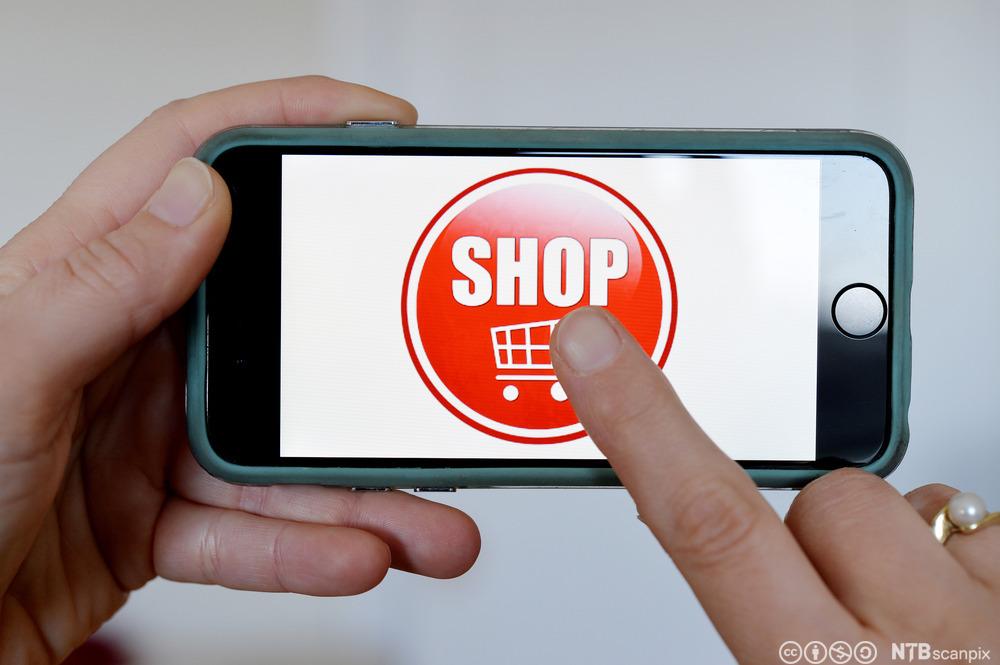 Mobiltelefon holdt av en person som skal til å klikke på en stor knapp på skjermen hvor det står "shop" og er bilde av en handlekurv. Foto.