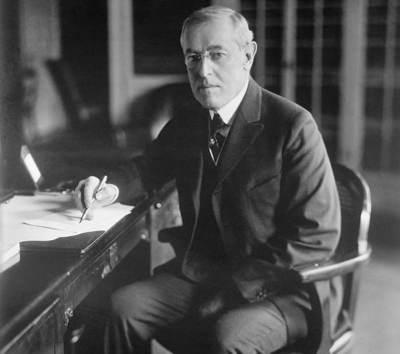 President Woodrow Wilson sit ved skrivebordet. Han har ein penn i høgre hand, og nokre papir ligg framfor han. Han ser mot fotografen. Foto.