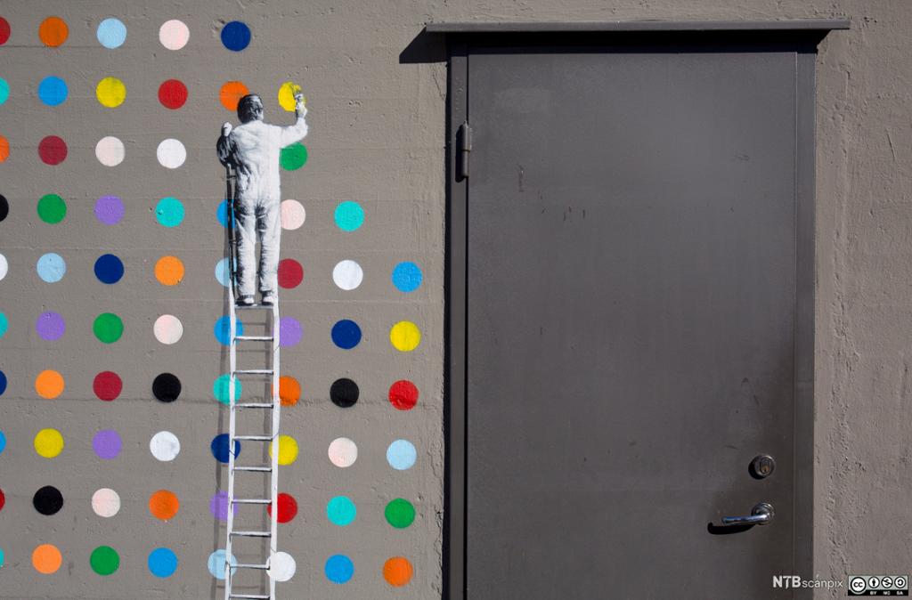 Veggmaleri som viser en mann på stige som maler fargeprikker. Foto.