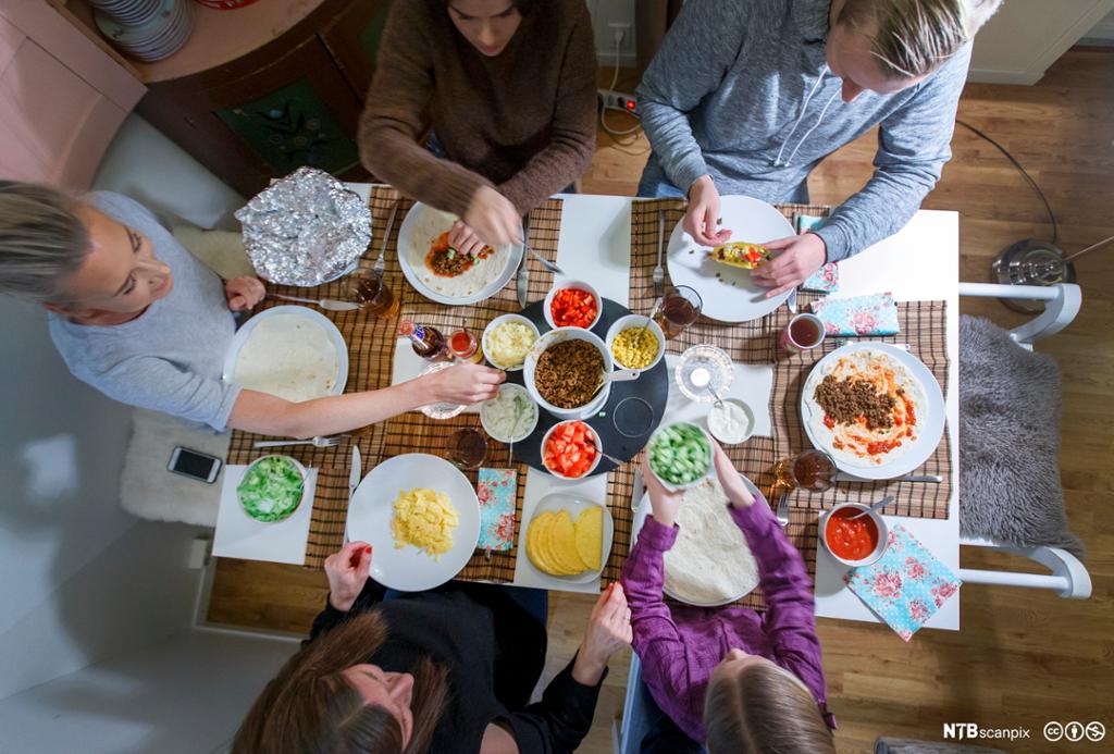 Fem personer sitter rundt et bord og spiser et tacomåltid sammen. Bildet er tatt rett ovenfra. Foto.