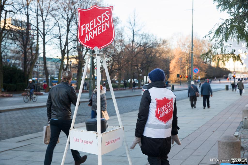 Julegryte fra Frelsesarmeen. Rundt gryta stimler folk forbi på gata. En person fra Frelsesarmeen står ved gryta. Hen har en hvit vest med rød logo. Foto.