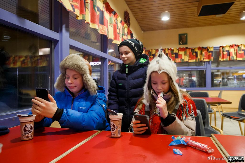 Tre barn spiser godteri og ser på mobiltelefonene sine. Foto.