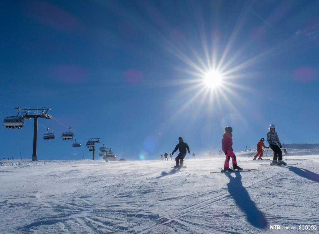 Foto av folk på ski og snøbrett i en slalåmbakke på en fin dag med strålende sol. 