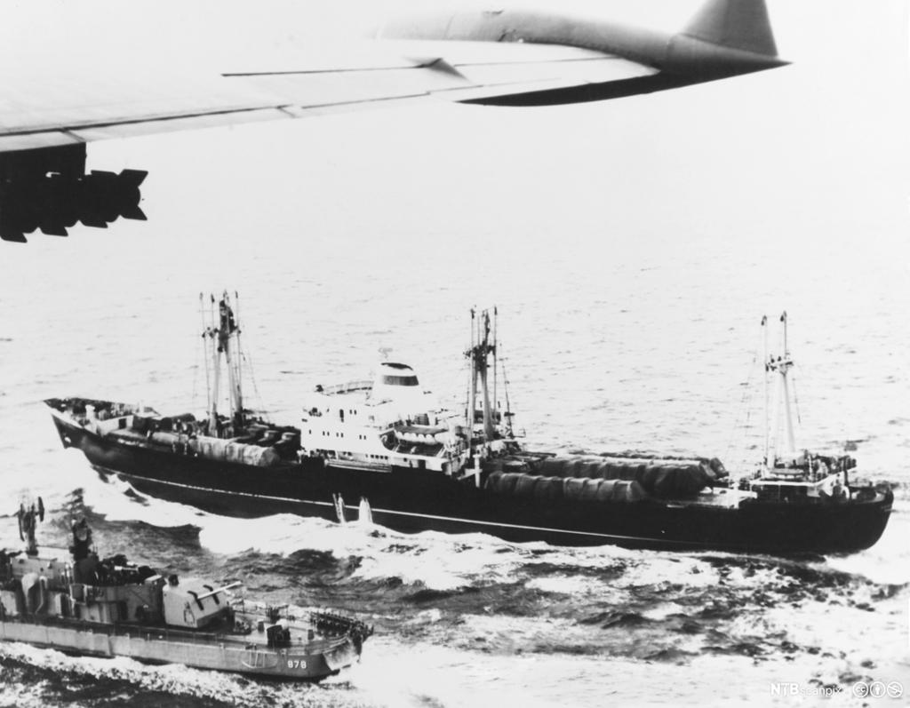 Flyfoto av eit skip lasta med svære rakettar. Ved sida av er det eit mindre marinefartøy. Det heng missil under flyvengen også. Foto.