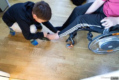 Gutt hjelper mann i rullestol med å ta på sko. Foto.