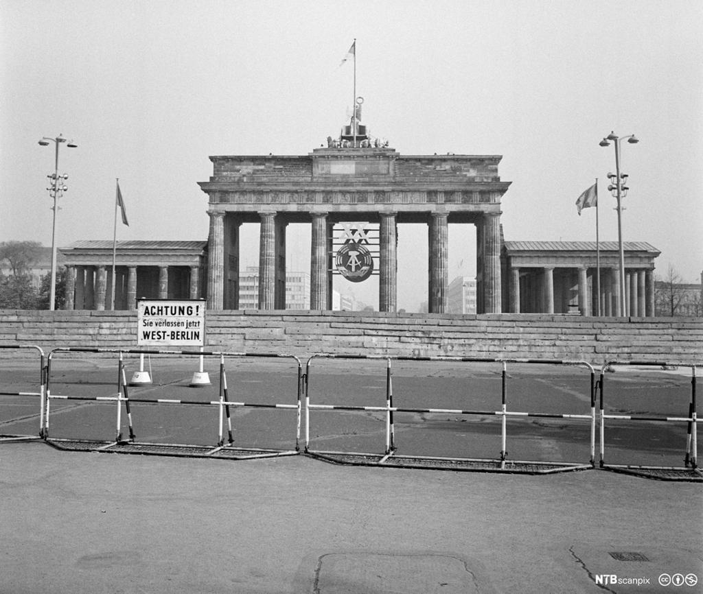 Brandenburger Tor fotografert frå Vest-Berlin i 1969. Fremst i biletet er det eit enkelt gjerde, bak det eit skilt der det på tysk står ei åtvaring om at ein forlet Vest-Berlin. Deretter ein høg mur framfor sjølve Brandenburger Tor. Inni det historiske monumentet er det hengt opp eit stort symbol for DDR-staten. Foto.