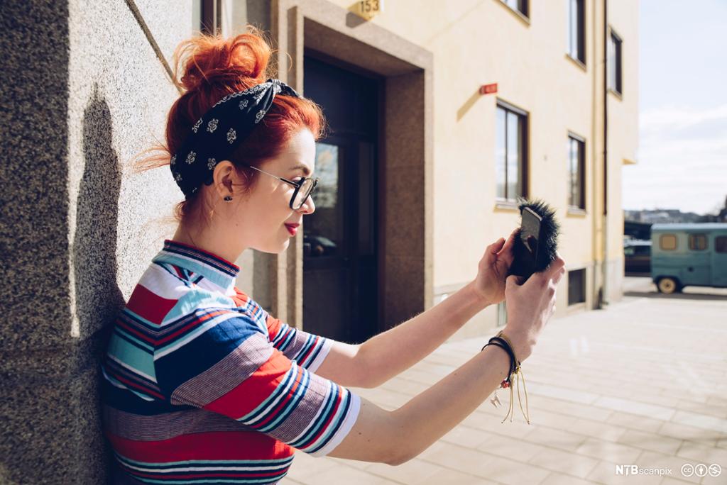 Ei jente med rødt hår, hårbånd og kortermet genser tar selfie mot en solvegg. Foto.