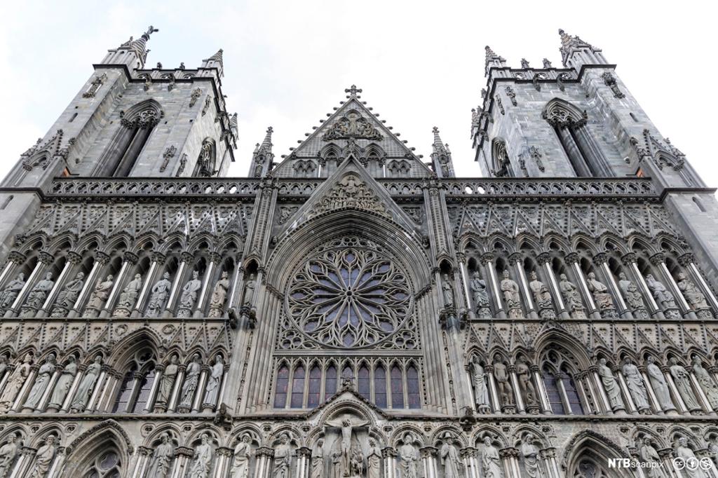 Fronten av katedral i gotisk stil med to tårn, helgenstatuar og rundt glasmåleri i sentrum. Foto.