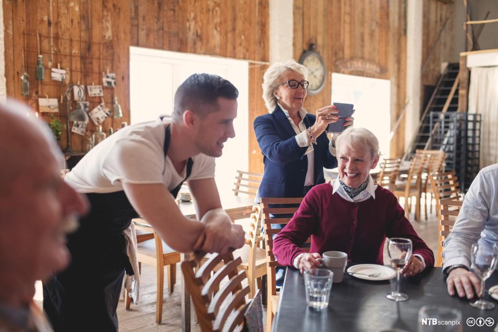 Ung servitør i uformell prat med eldre gjester. Foto.