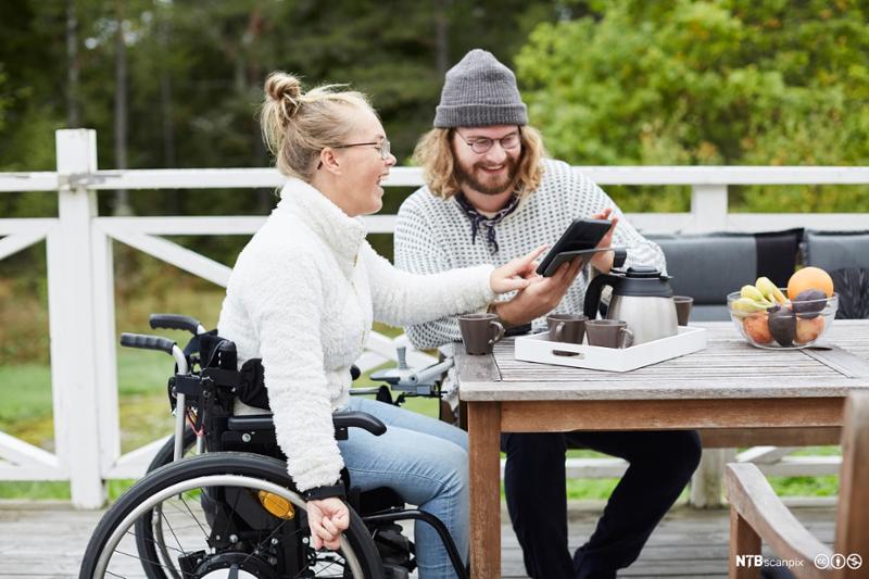 Ung kvinne i rullestol sitter ved hagebord sammen med ung mann. De ser på en skjerm sammen og ler. Foto.
