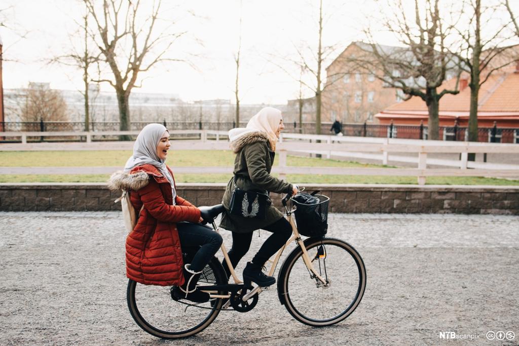 To unge kvinner med hijab på felles sykkel. Foto.