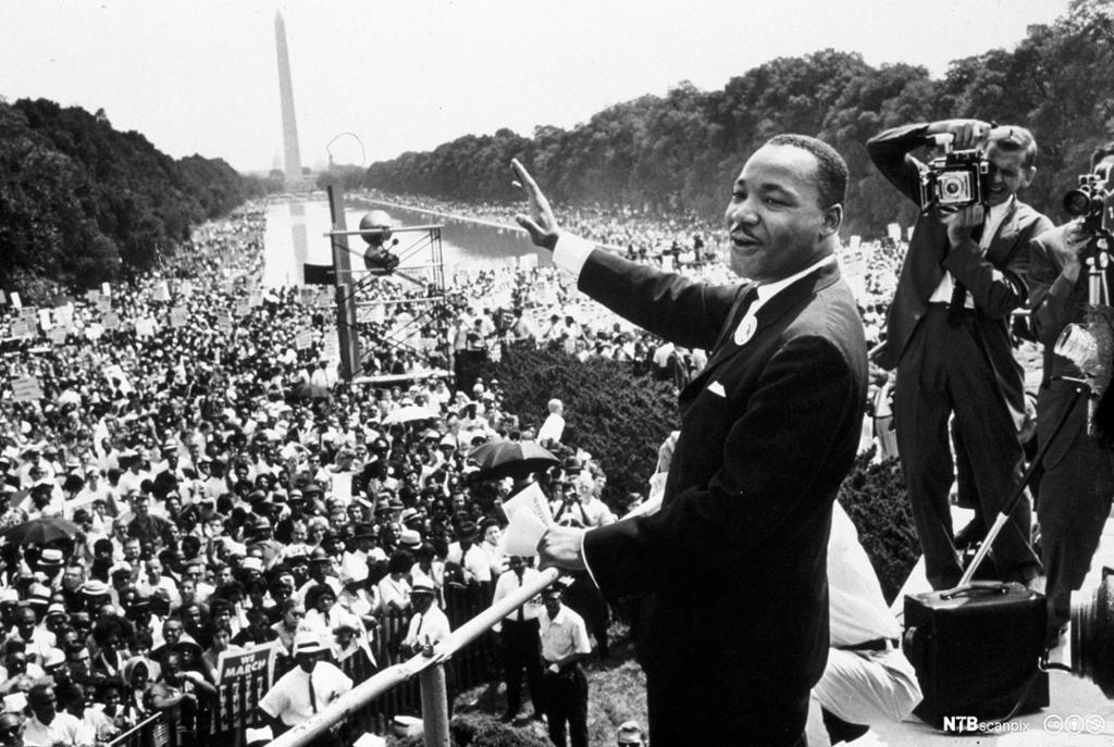 Photo: We see Martin Luther King Jr. waving at a large crowd of people. Behind him a photographer is taking his picture. In the background we see the Washington monument. 