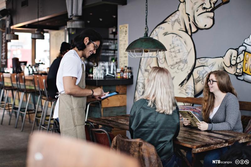 En ung servitør på en kafé tar imot bestillingen fra to gjester. Både han og gjestene smiler. Foto.
