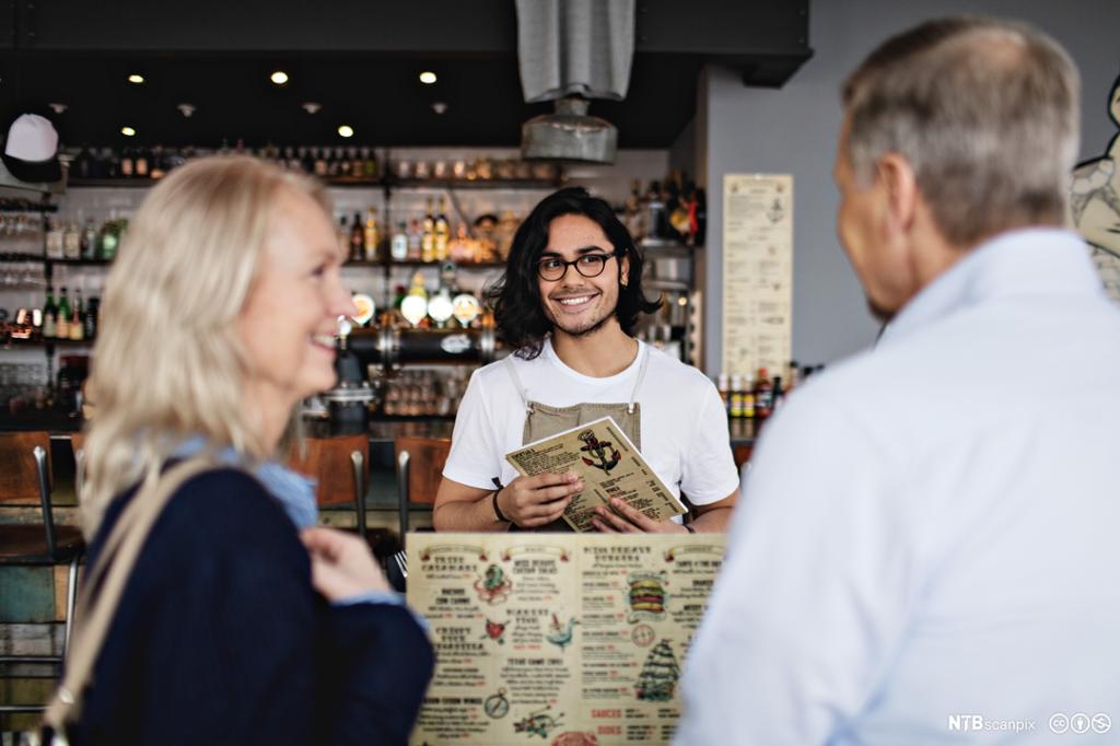 En smilende servitør snakker med en mann og en dame i en restaurant. Foto.