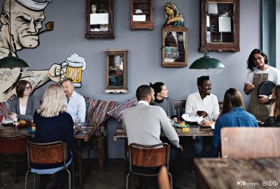 Servitør snakker med gjestene ved et av bordene i en kafe. Foto.