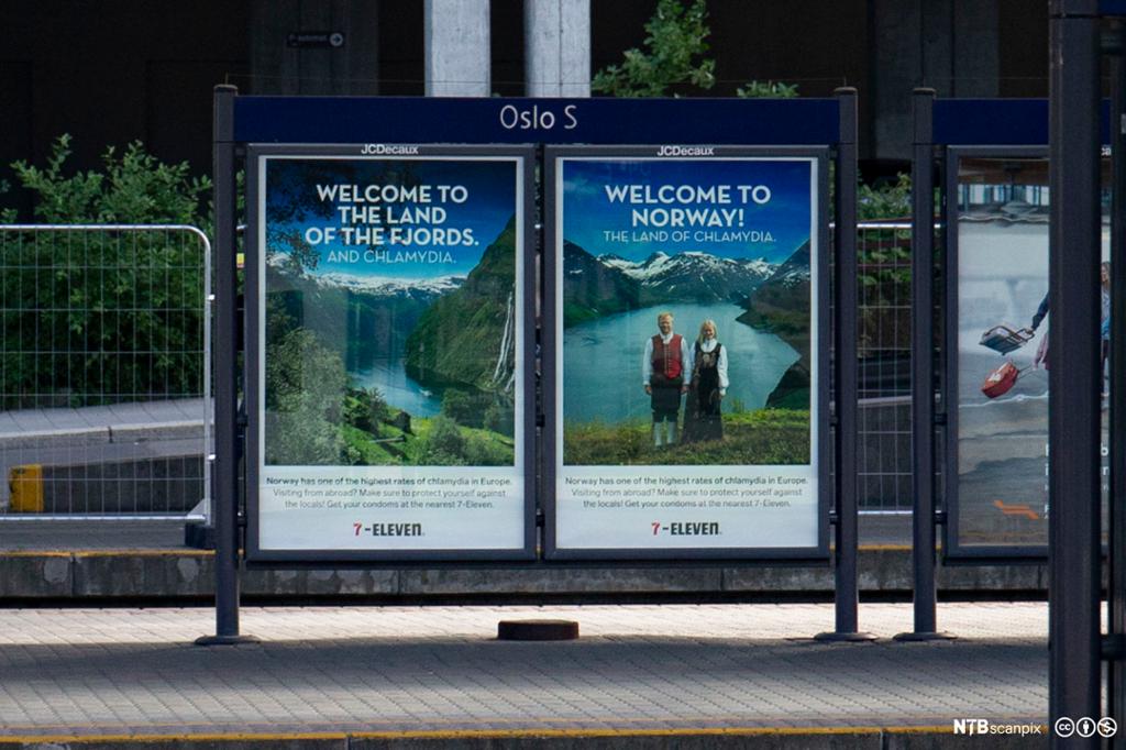 To reklameplakater for 7-Eleven er satt opp på en togplattform på Oslo S. De består av flotte bilder av fjord, fjell og et blondt par i bunad, og en tekst som sier «Welcome to Norway – the land of Chlamydia».  Foto.