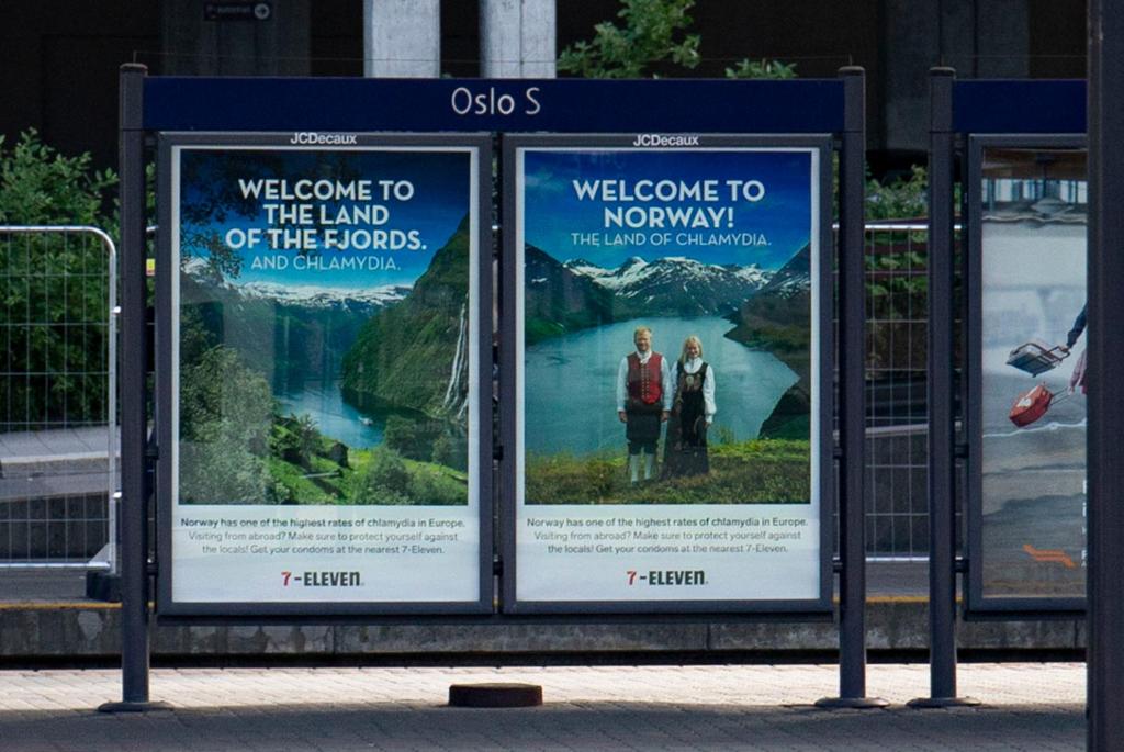 To reklameplakater for 7-Eleven er satt opp på en togplattform på Oslo S. De består av flotte bilder av fjord, fjell og et blondt par i bunad og en tekst som sier "Welcome to Norway – the land of Chlamydia".  Foto.