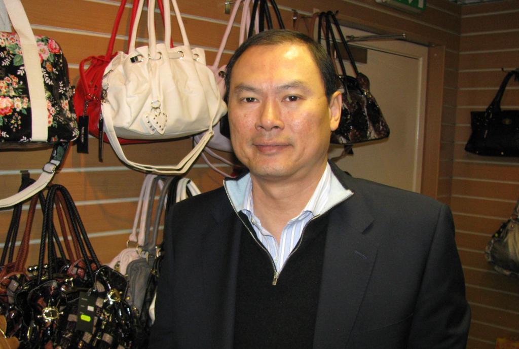 Photo: Adult Asian man who is wearing a suit jacket, sweater, and open shirt. He is looking at the camera. He appears to be in a shop that sells handbags. 