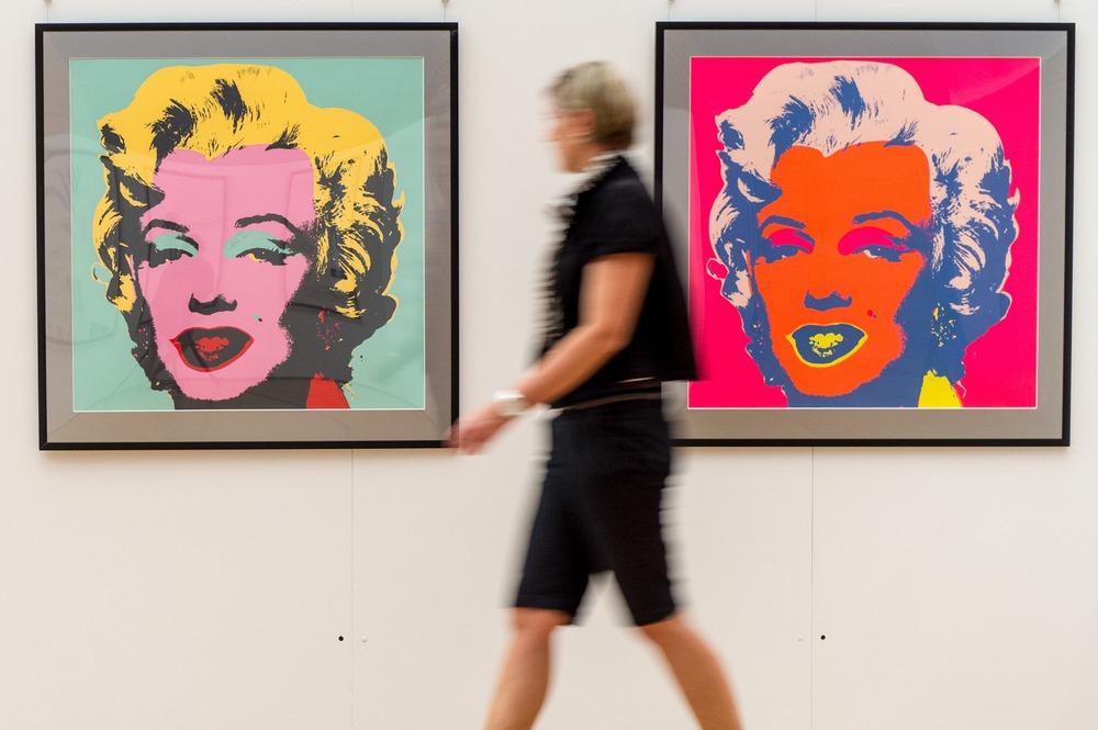 En kvinne går forbi en vegg med to fargerike, innramma illustrasjoner av et kvinneansikt. Bildene på veggen er to av Andy Warhols portretter av Marilyn Monroe. Foto.