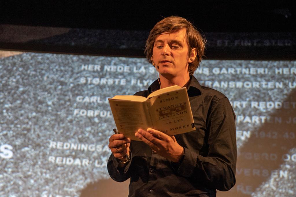 Mann med mikrofon står på ein scene og les frå ei bok med tittelen "Leksikon om lys og mørke". Foto.