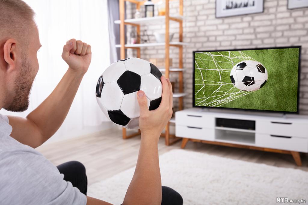 En mann sitter i sofaen med en fotball og ser fotball på TV. Foto.