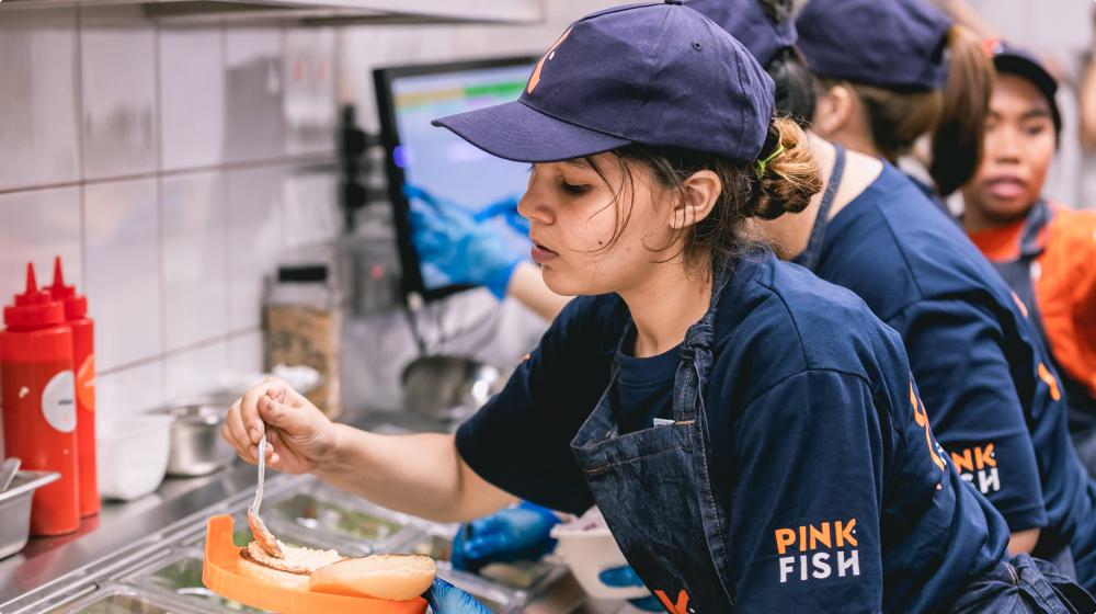 En Pink Fish-ansatt ordner med en lakseburger. Hun har mørkeblå klær og kaps med Pink Fish-logo på. Flere andre ansatte står i bakgrunnen. Foto.