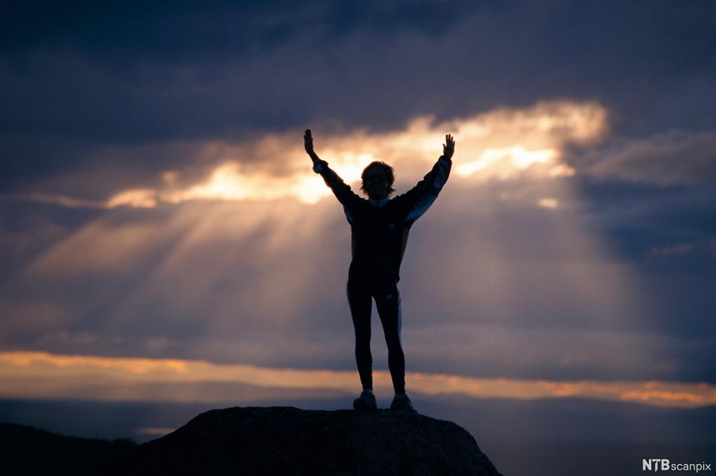 En person strekker armene opp mot en himmel der sola bryter gjennom skyene. Vi ser personen bakfra, og hen står på en fjelltopp og ser utover. Foto.