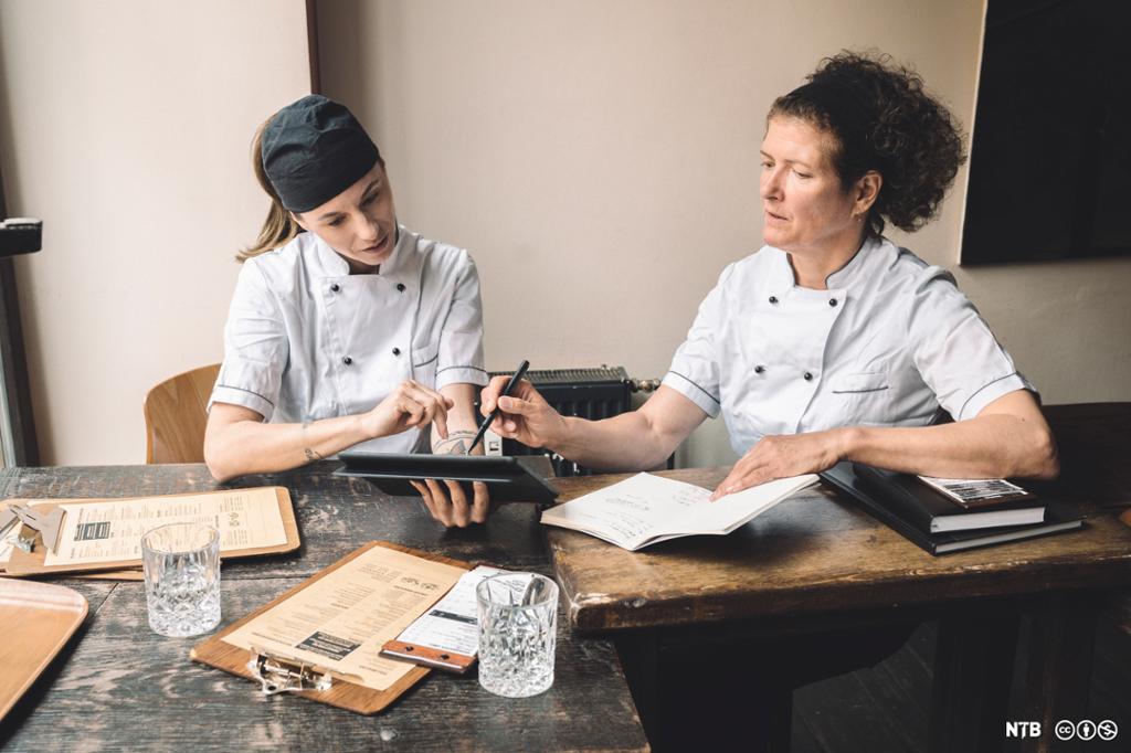 Foto av to kokkar som sit ved eit bord og diskuterer, med ein iPad, menyar og skrivebøker framfor seg.