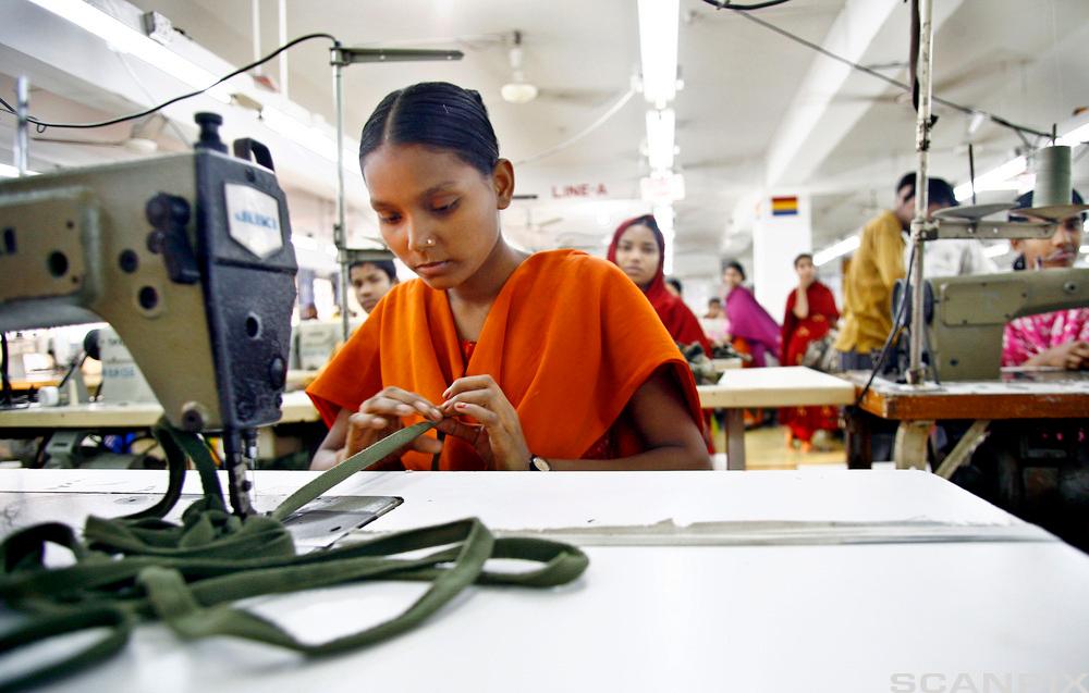 Ung jente sit ved ein symaskin på ein klesfabrikk i Bangladesh. I bakgrunnen er fleire tekstilarbeidarar som sit ved symaskinene, alle i fargerike klede. Foto.