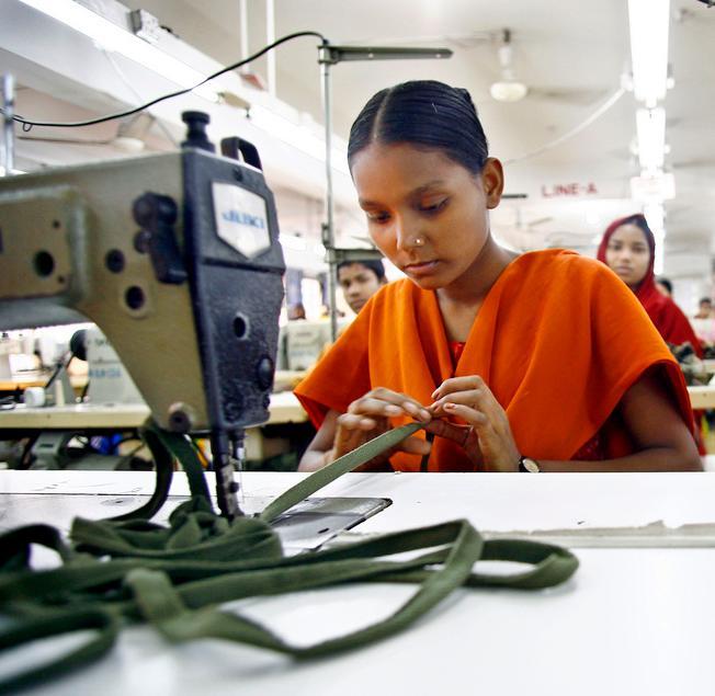 Tekstilarbeidar i Bangladesh: Ung kvinne i oransje kjole sit ved ei symaskin og arbeider. I bakgrunnen er to andre arbeidarar. Foto.