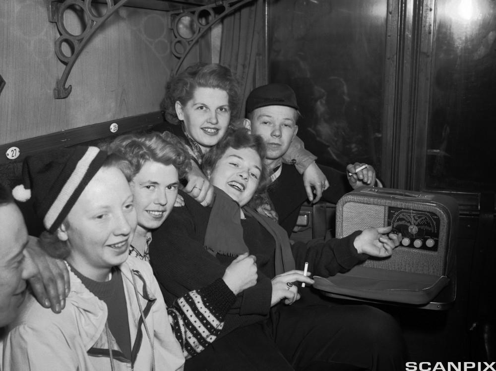En rekke ungdommer på et gammelt svarthvitt bilde. De har det gøy og røyker sigaretter. Ei jente skrur på en radio. Foto.