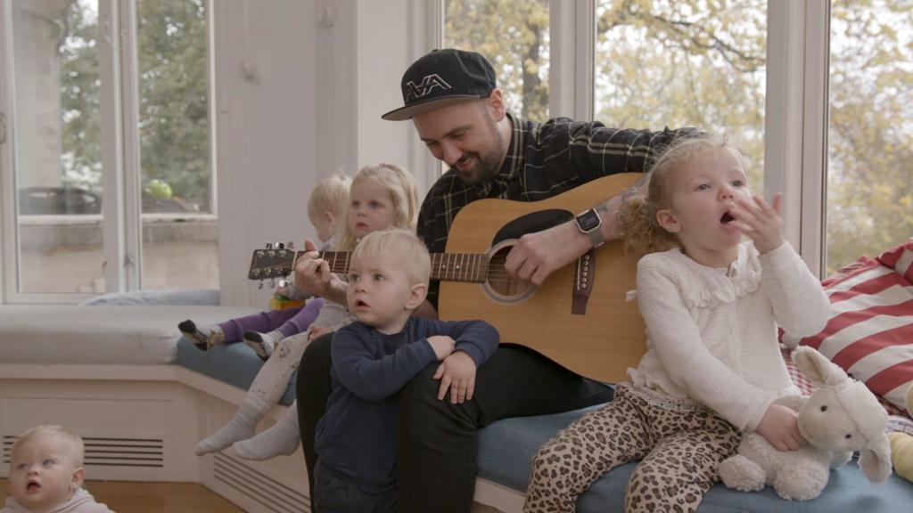 Ein mannleg barnehagetilsett med gitar og fleire små barn rundt seg. Foto.