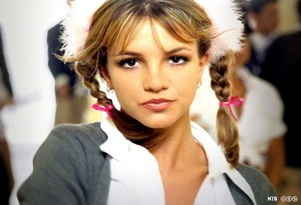 En ung Britney Spears ser alvorlig inn i kameraet. Hun har musefletter og grå skoleuniform. Stillbilde fra musikkvideoen «Baby One More Time».