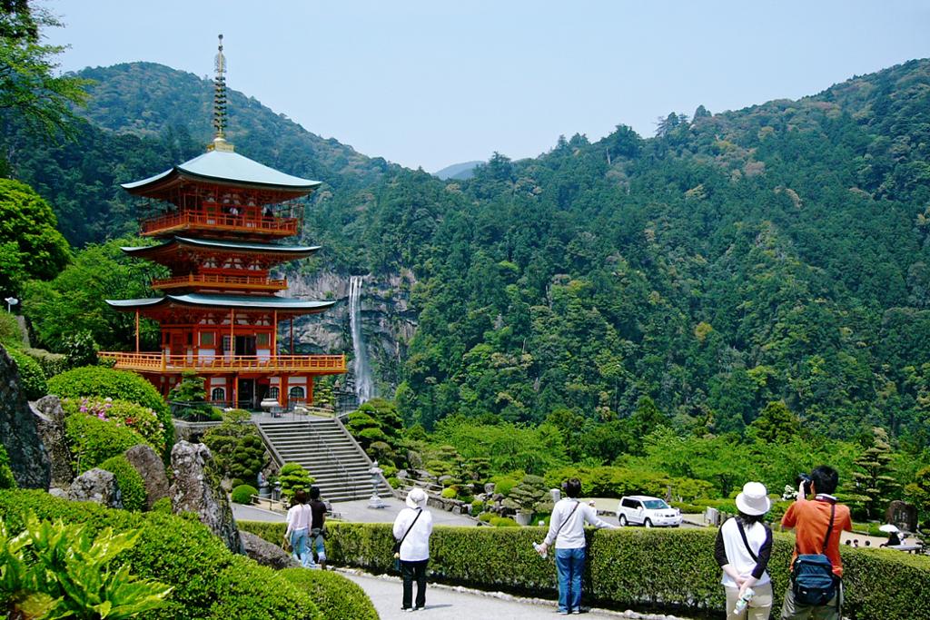 Treetasjes pagode-tempel med foss i bakgrunnen. Nokre personar står og ser på og beundrar tempelet. Ein person fotograferer tempelet. Foto.