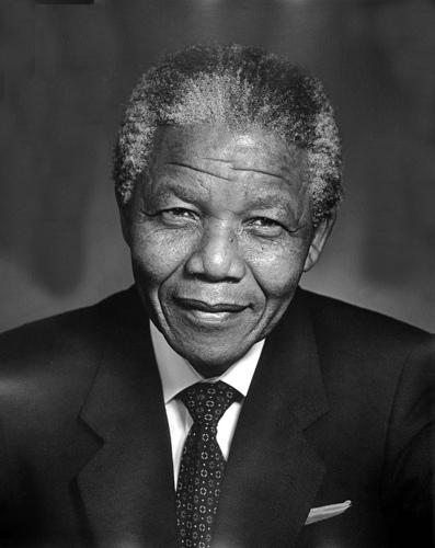 A black and white photo of Nelson Mandela. He is an old man. He's dressed in a suit and tie. He looks directly into the camera, smiling.  