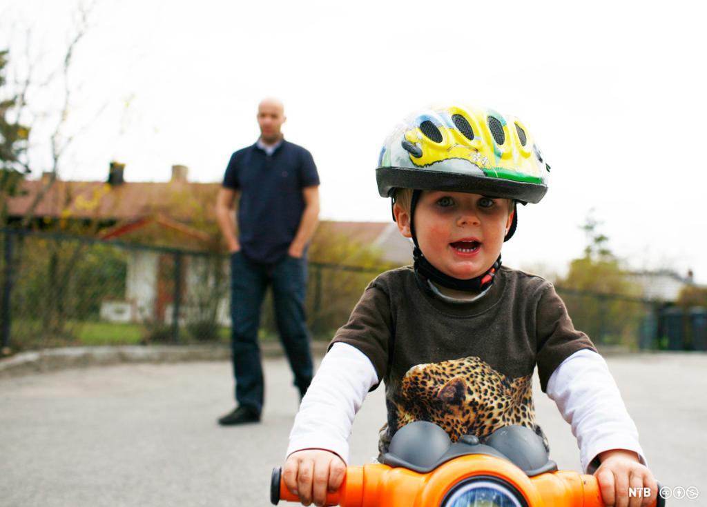 Ein liten gut med sykkelhjelm syklar konsentrert. I bakgrunnen står ein mann og følgjer med. Foto.