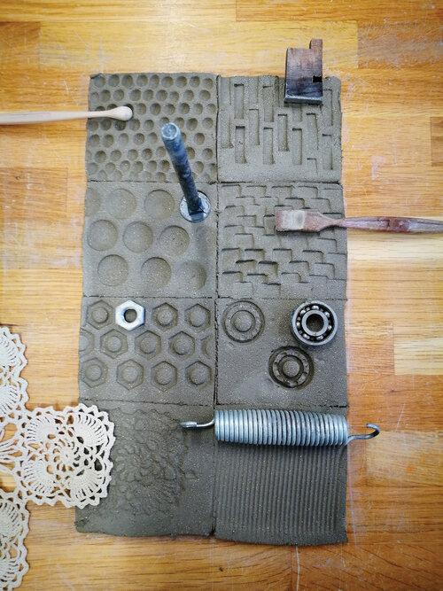Ulike gjenstander som skruer og en heklet duk som lager avtrykk i leire, ligger på ei bordplate. Foto.