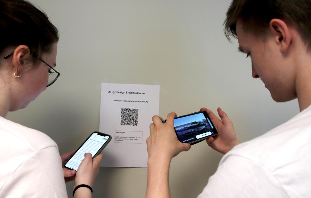 To elevar står ved eit ark med ein QR-kode og skannar koden med kvar sin mobil. Foto.