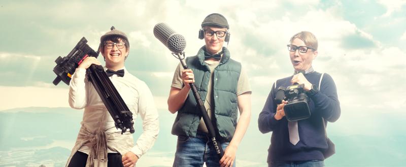 Tre gutter med briller smiler mot kamera.  De bærer på videokameraer og en mikrofonbom. Foto.