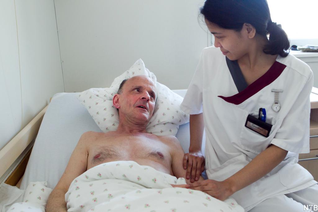Helsefagarbeider viser omsorg for sengeliggende pasient ved å sitte på sengekanten, holde ham på armen, smile og ha øyekontakt når han snakker til henne. Foto.