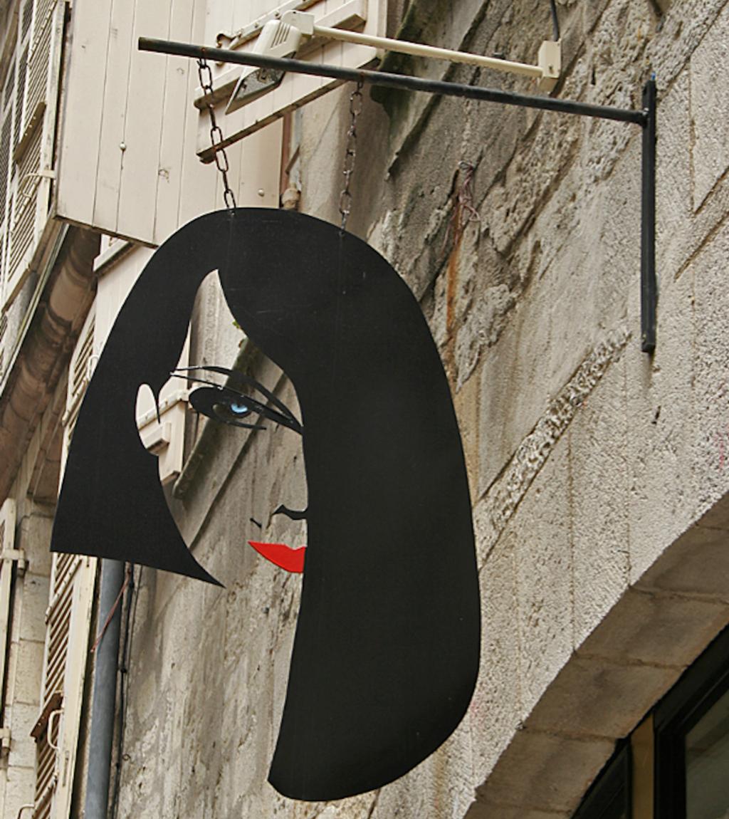 Fotografi av skilt som viser en kvinne med svart hår og røde lepper. Skiltet henger ut fra en vegg.